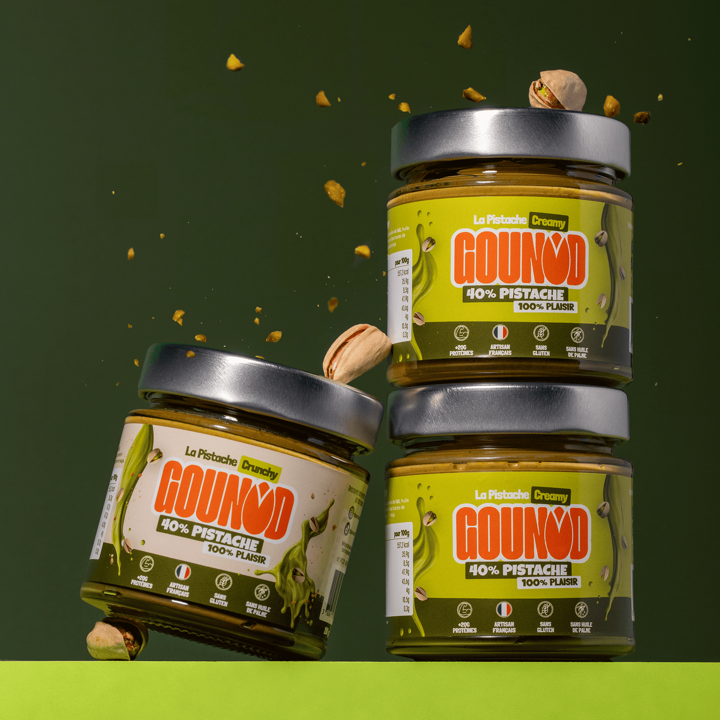 Pots de pâte à tartiner pistache Gounod empilés, versions Creamy et Crunchy, avec éclats de pistache en suspension.