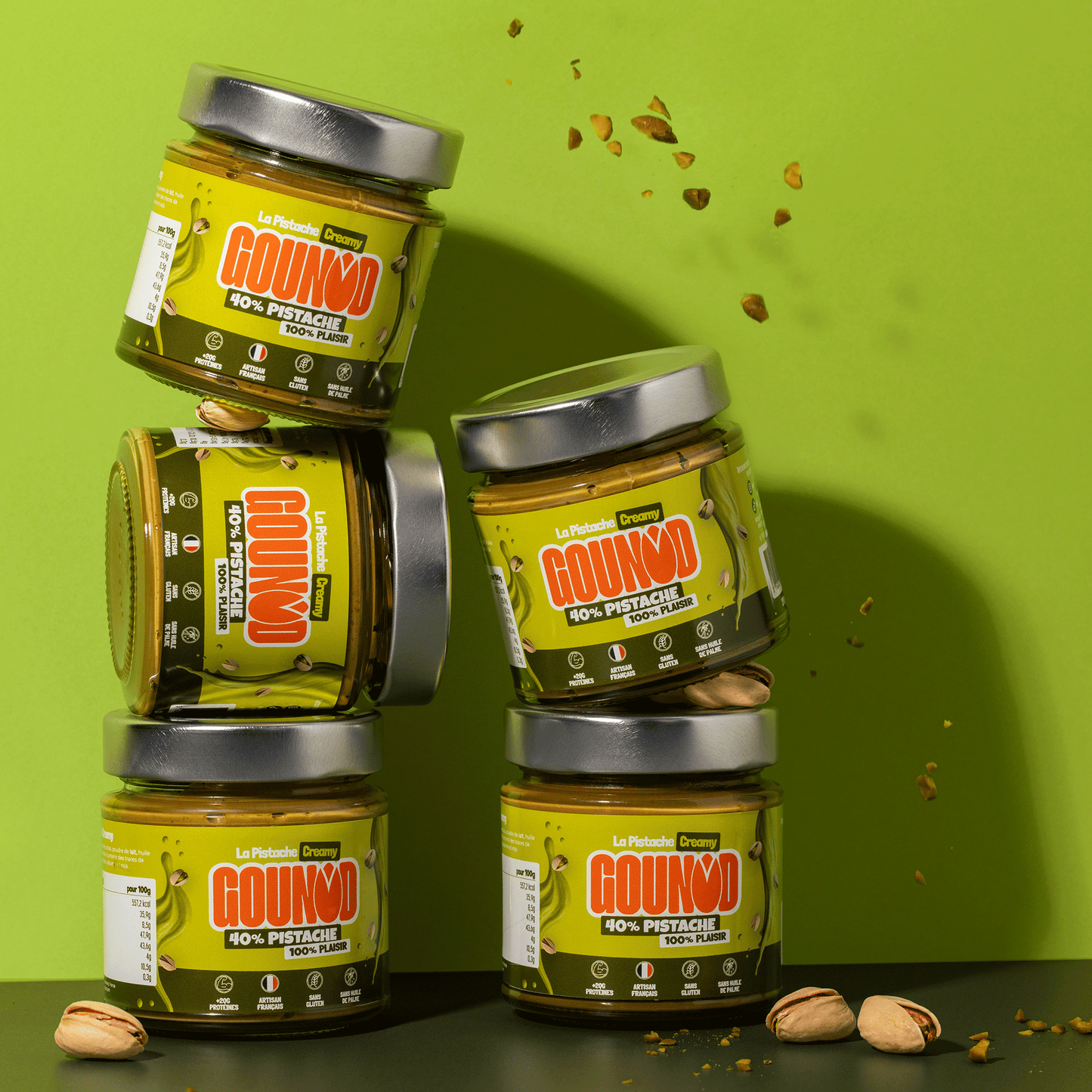 Empilement de 5 pots de pâte à tartiner pistache Gounod Creamy sur fond vert, avec éclats de pistache en suspension.