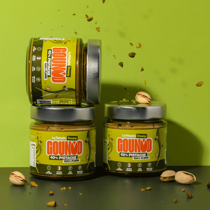 Pots de pâte à tartiner pistache Gounod Creamy empilés avec des éclats et des pistaches tombant sur fond vert.