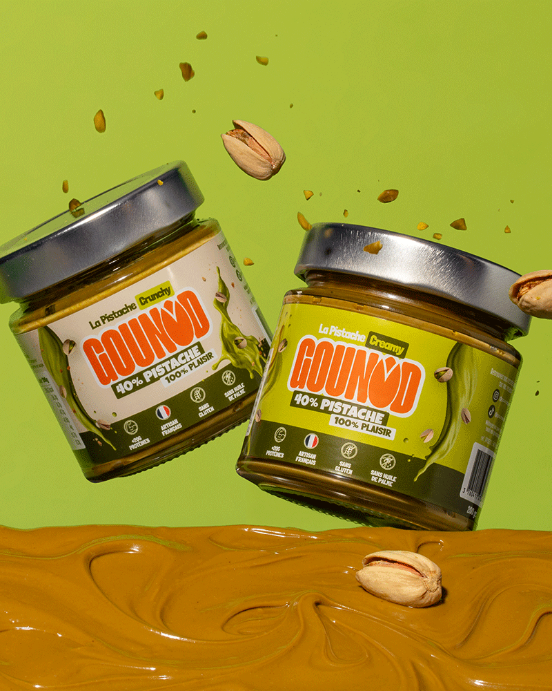 Deux pots de pâte à tartiner pistache Gounod, Creamy et Crunchy, en suspension au-dessus d’un fond vert avec éclats de pistache et pâte à tartiner en dessous.