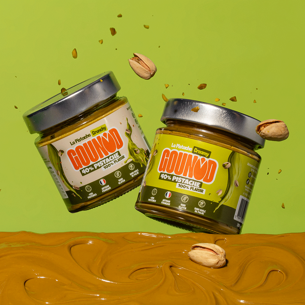 Deux pots de pâte à tartiner pistache Gounod, Creamy et Crunchy, en suspension au-dessus d’un fond vert avec éclats de pistache et pâte à tartiner en dessous.