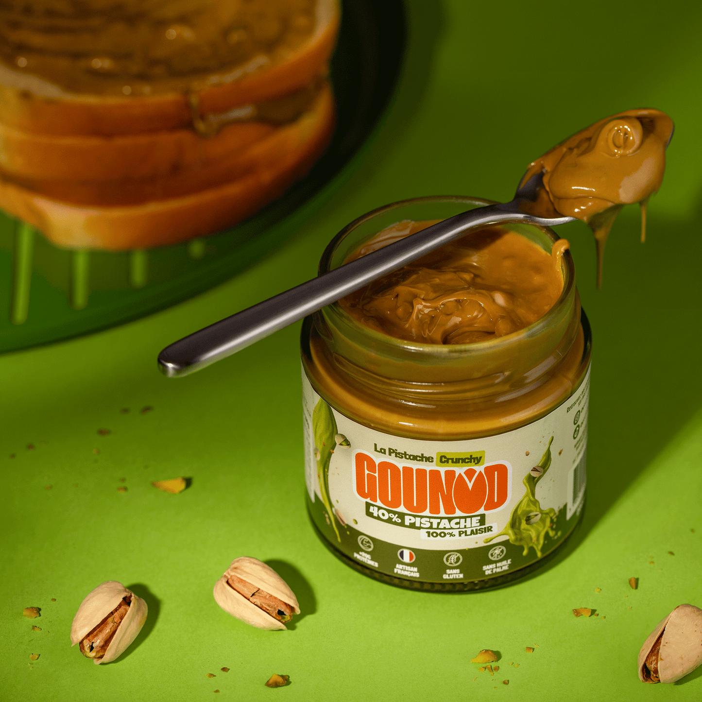 Pot de pâte à tartiner pistache Gounod Crunchy ouvert avec une cuillère recouverte de pâte, entouré de pistaches sur fond vert.