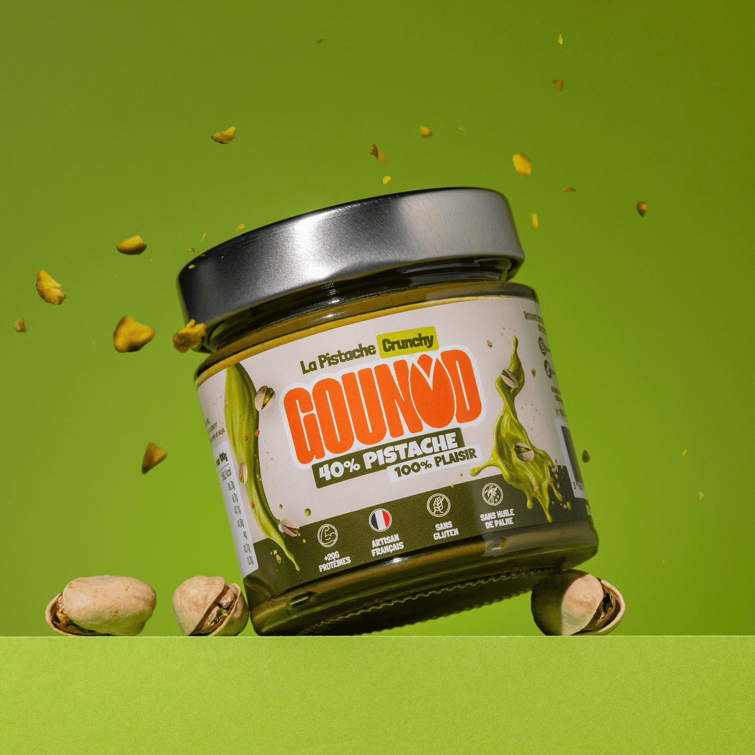 Pot de pâte à tartiner pistache Gounod Crunchy incliné, entouré d’éclats et de pistaches entières sur fond vert.